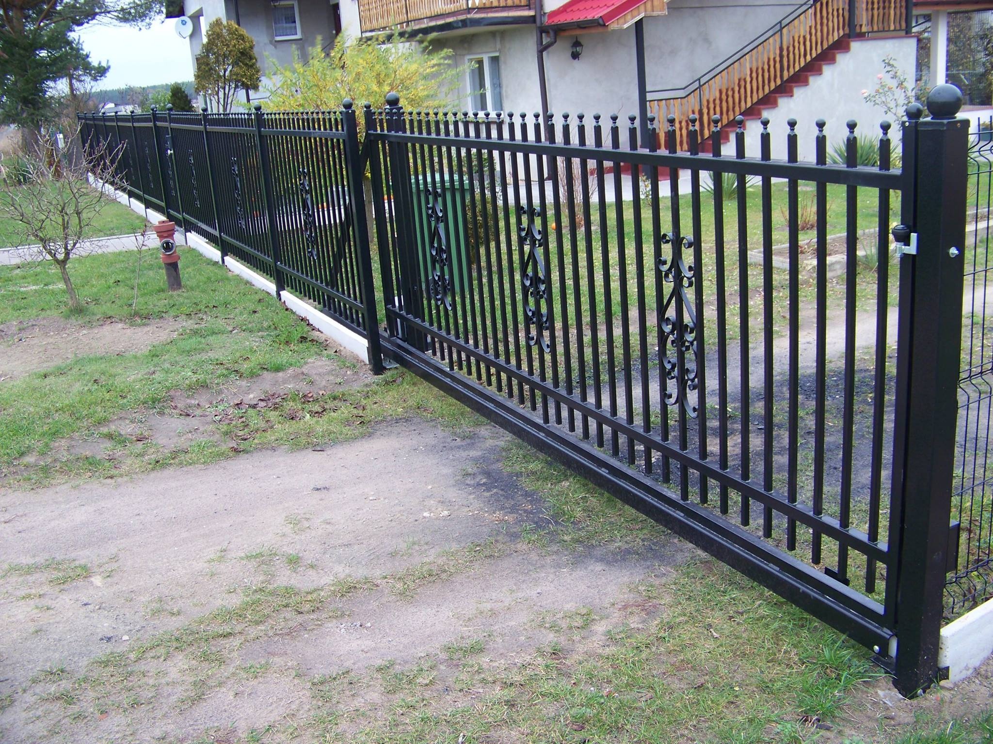 Czarna brama wjazdowa przesuwna z metalowymi prętami i ozdobnymi elementami, widoczna w perspektywie z boku na tle domu z czerwonym dachem i zielenią.