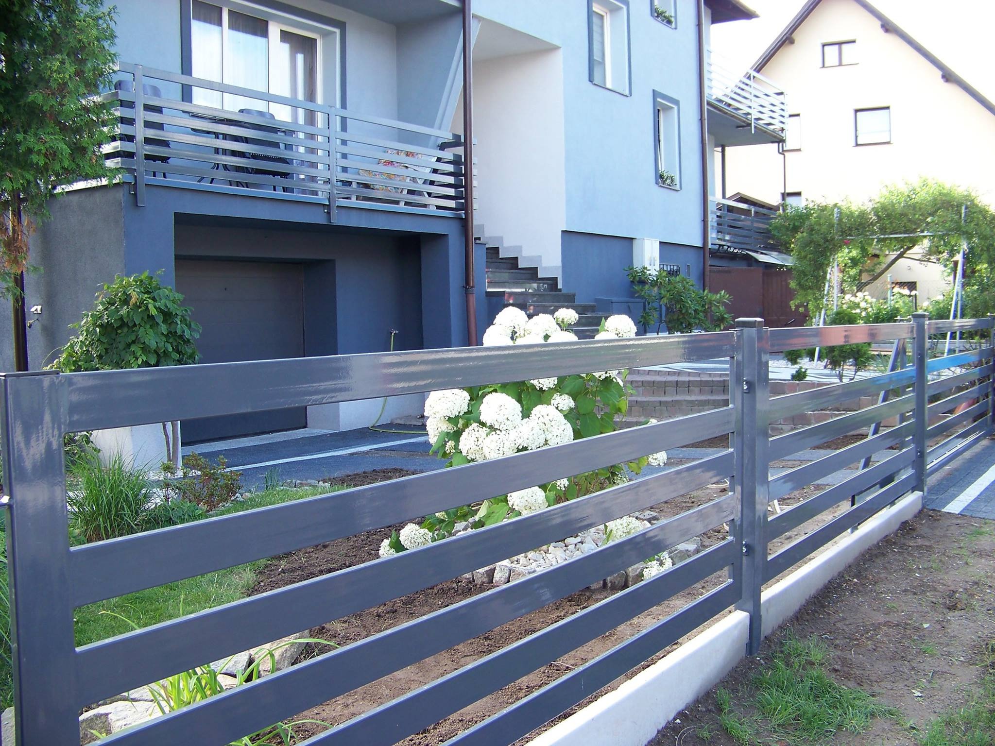 Nowoczesne, poziome ogrodzenie metalowe w kolorze grafitowym, otaczające posesję z domem w tle, z widocznymi białymi hortensjami.