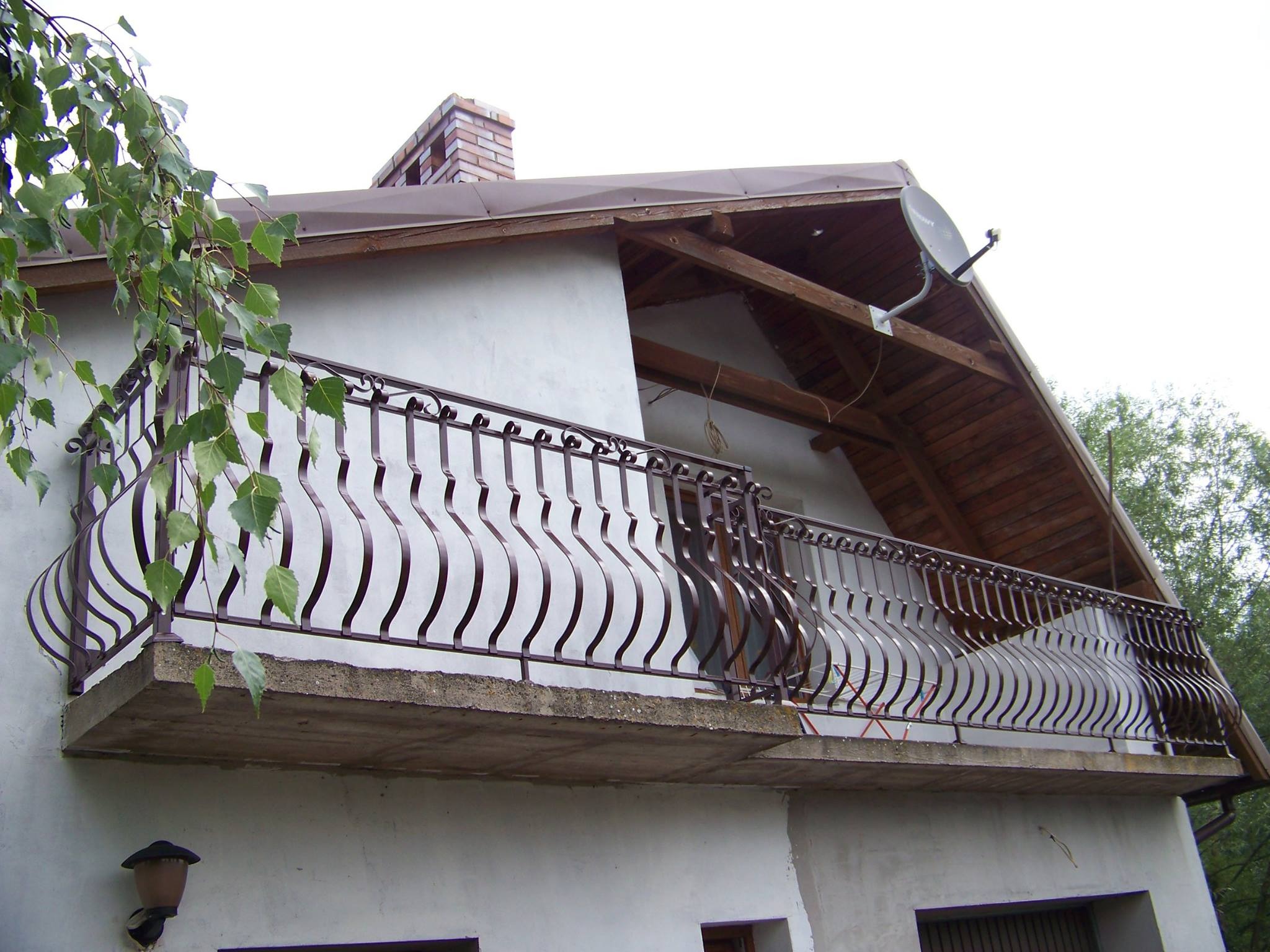 Balkon z metalową balustradą o falistym wzorze, widok z dołu, z fragmentem zwisających gałęzi drzewa w lewym górnym rogu, na tle białej ściany budynku z brązowym dachem i kominem.