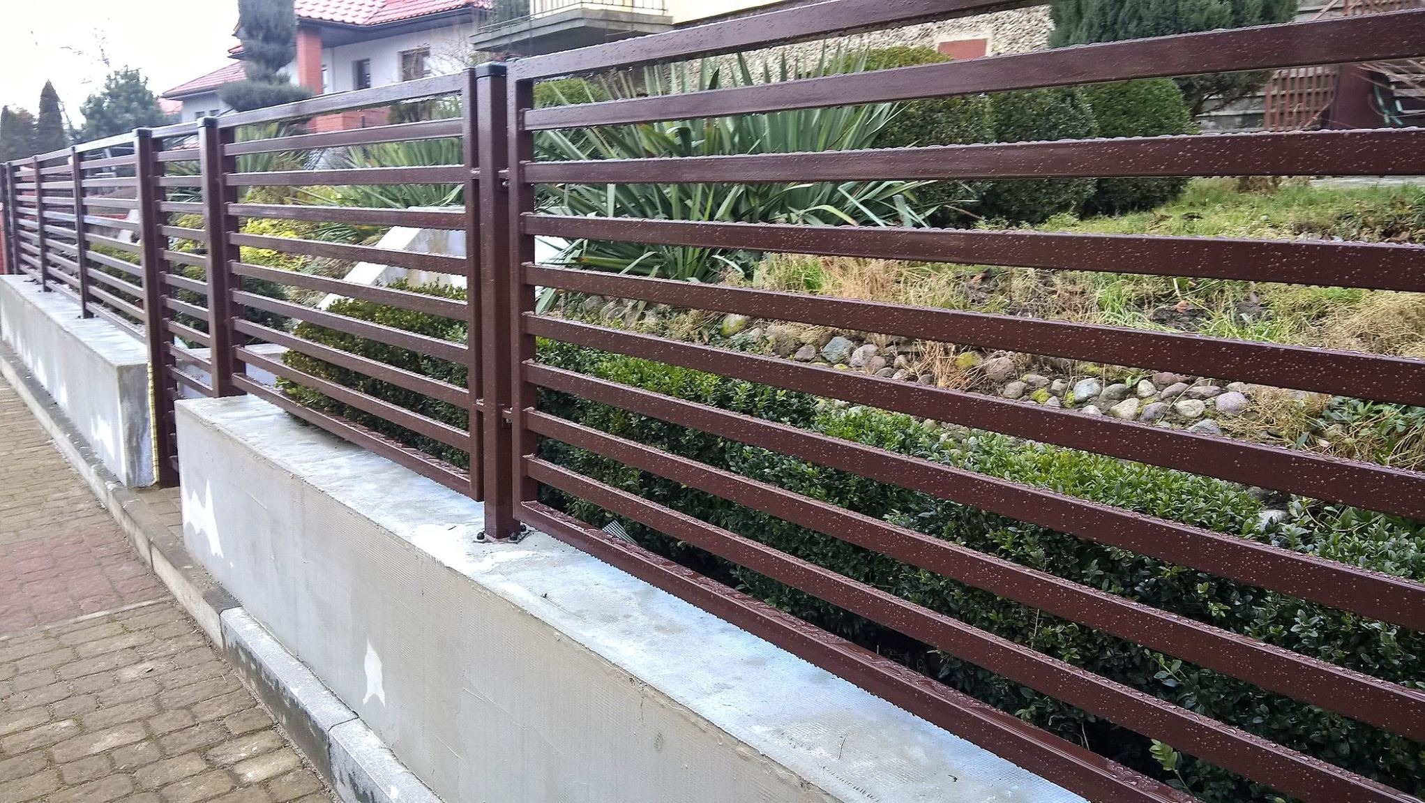 Nowoczesne ogrodzenie metalowe w kolorze brązowym, z poziomymi przęsłami, zamontowane na betonowym murku, widok z poziomu chodnika.