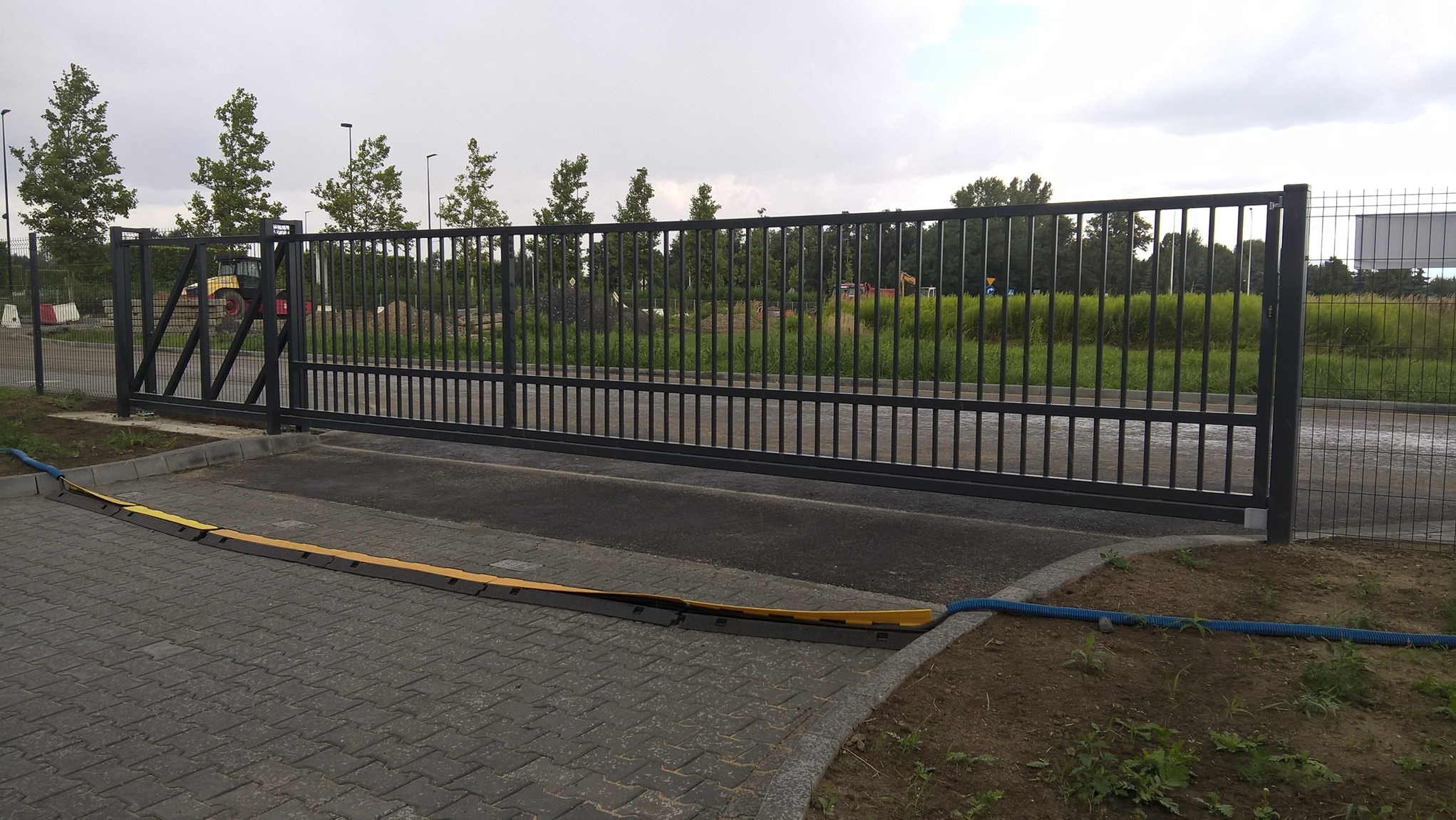 Przesuwna brama przemysłowa z czarnego metalu z zabezpieczeniem najazdowym na kostce brukowej; w tle teren budowy z widoczną koparką.