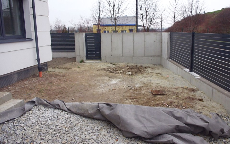 Przygotowany teren pod przyszły ogród: wyrównana ziemia, częściowo odsłonięte kamienie, szara plandeka przykrywająca żwir, betonowe ogrodzenie z metalową furtką, widok z boku domu.