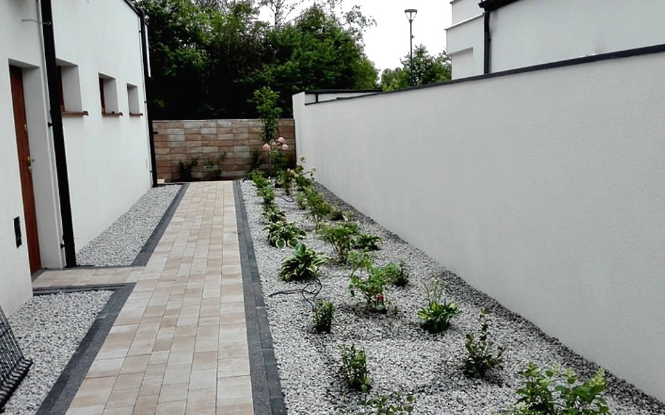 Ścieżka z kostki brukowej obłożona drobnym kamieniem prowadząca wzdłuż białego muru i budynku z nasadzeniami roślinnymi po obu stronach.