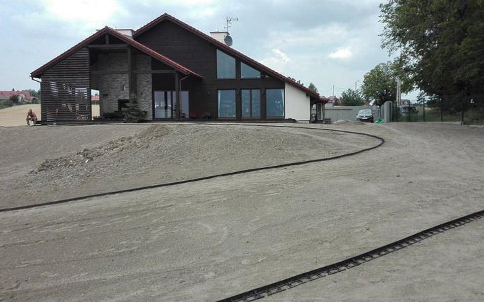 Przygotowanie terenu pod ogród przed nowoczesnym domem z dużymi oknami, widoczny wyrównany teren z czarną rurą nawadniającą i siedzący mężczyzna w oddali.