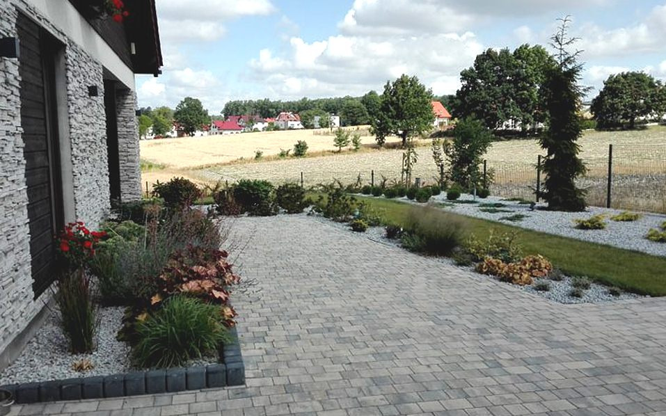 Ułożona kostka brukowa przed domem z kamienną elewacją, widoczne rabaty z różnorodną roślinnością obsypane białym żwirem, w tle pole i zabudowania.