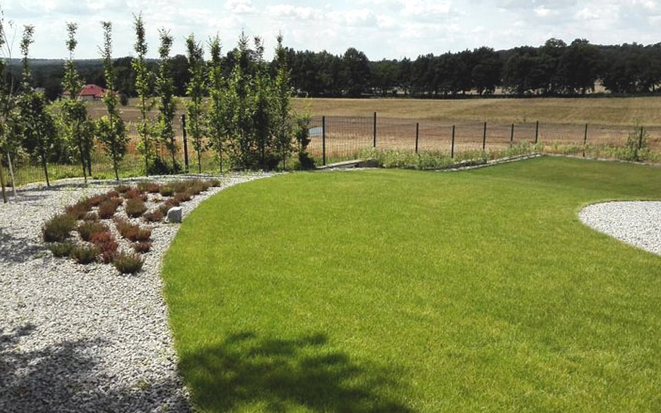 Świeżo założony trawnik z rolki, obrzeża z białego grysu, rabata z niskich krzewów i młode drzewka wzdłuż ogrodzenia, widok na pola i las w tle.