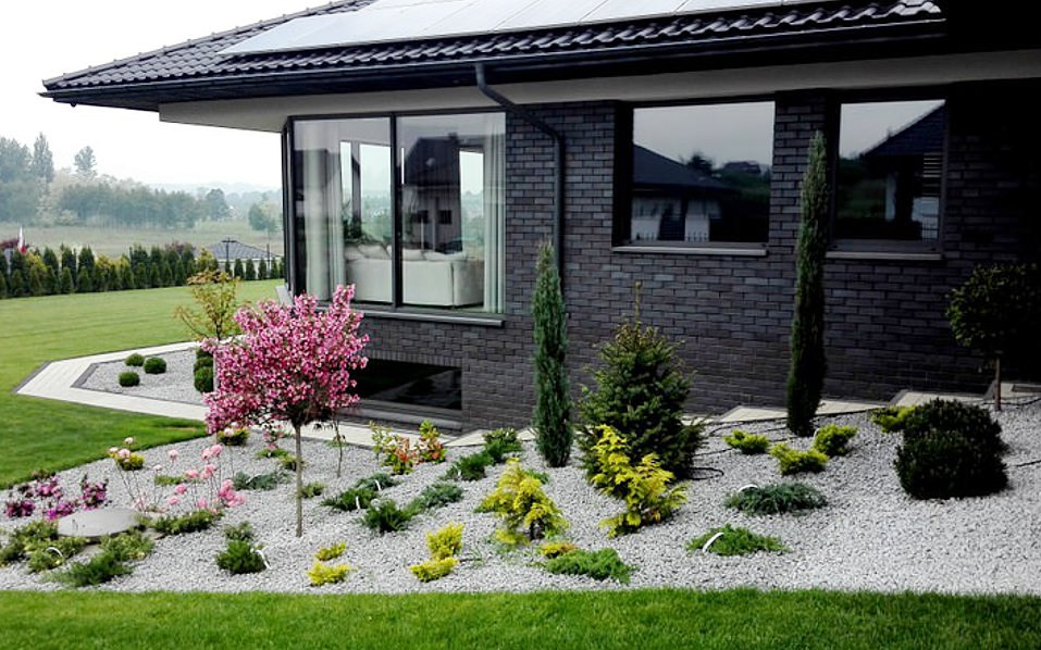 Nowoczesny ogród przydomowy z kwitnącym drzewem wiśniowym, ozdobnymi krzewami i wysypanym białym kamieniem, na tle ciemnej cegły domu z dużymi oknami.