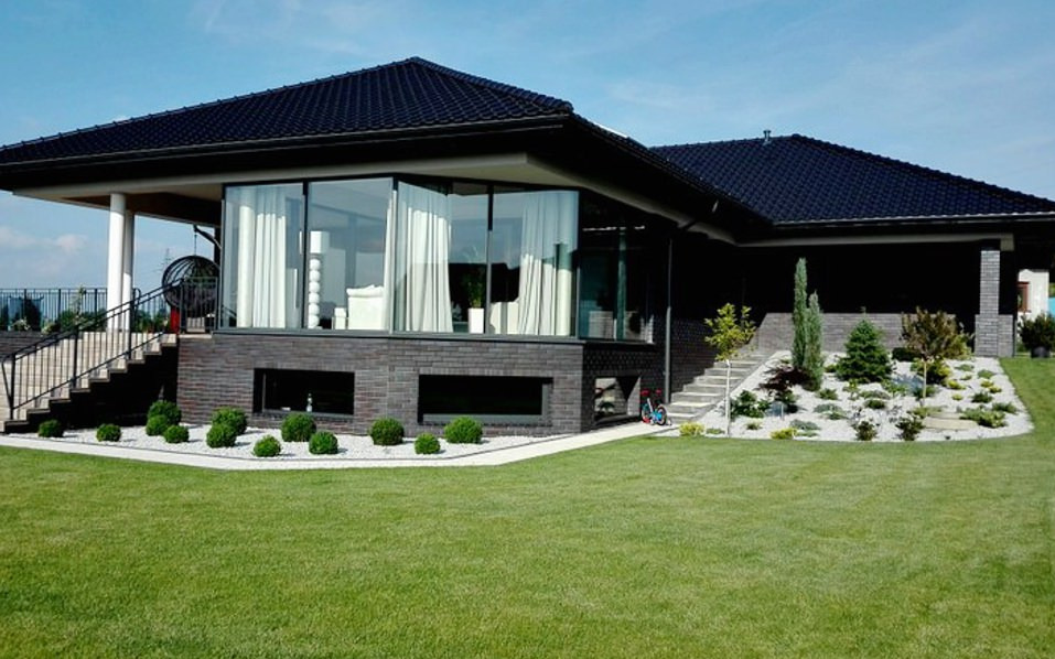 Nowoczesny dom z dużymi oknami i eleganckim ogrodem z równo przystrzyżonym trawnikiem, rabatami z białym kamieniem i niskimi krzewami.