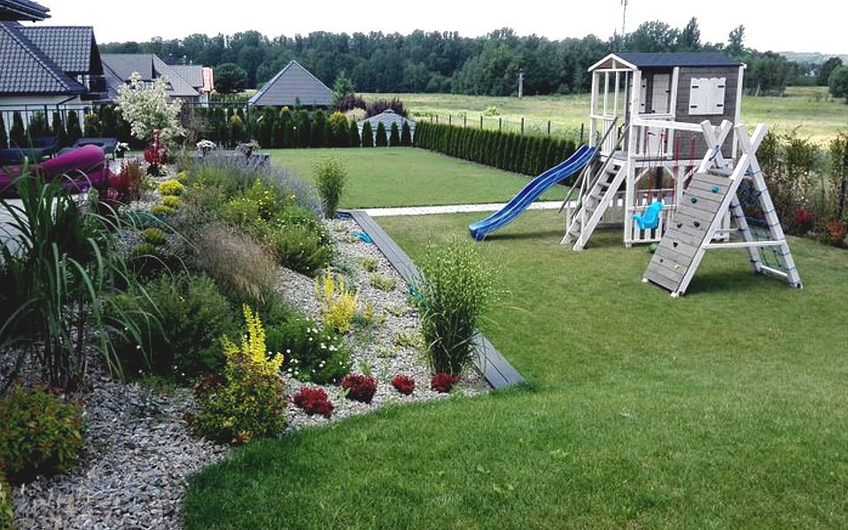 Ujęcie ogrodu z placem zabaw, rabatą kwiatową z kamieniami i tujami w tle, w słoneczny dzień.