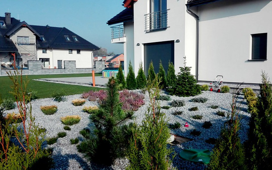 Ogród przydomowy z nasadzeniami iglaków, wrzosów i krzewów ozdobnych na tle białego domu z ciemną bramą garażową, wysypany jasnym grysem.