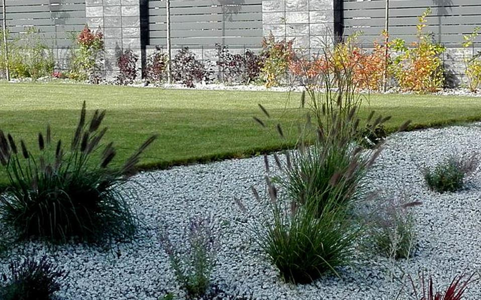 Nowoczesny ogród z ozdobnymi trawami i białym żwirem, widok na zadbany trawnik i szary płot panelowy.