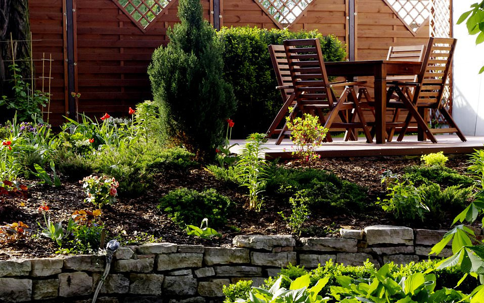 Ogród z kamienną skarpą, roślinami, tujami i drewnianym zestawem mebli ogrodowych na tarasie z desek, w tle drewniany płot z kratą.