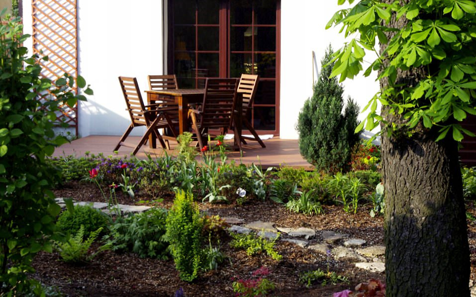 Aranżacja ogrodu z drewnianym tarasem, meblami ogrodowymi i ścieżką z kamieni, widok na dom z brązowymi drzwiami.