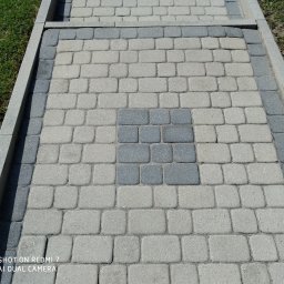 Stop Plamom - Ułożona kostka brukowa w odcieniach szarości z ciemniejszym kwadratem pośrodku. Widoczne krawężniki i trawa. Zdjęcie z bliska, ukazujące strukturę kostki.