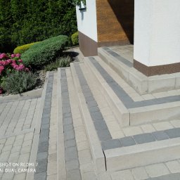 Stop Plamom - Schody i chodnik z kostki brukowej w odcieniach szarości i beżu, prowadzące do budynku z elementami drewnianymi i kolumną. Obok rosną hortensje.