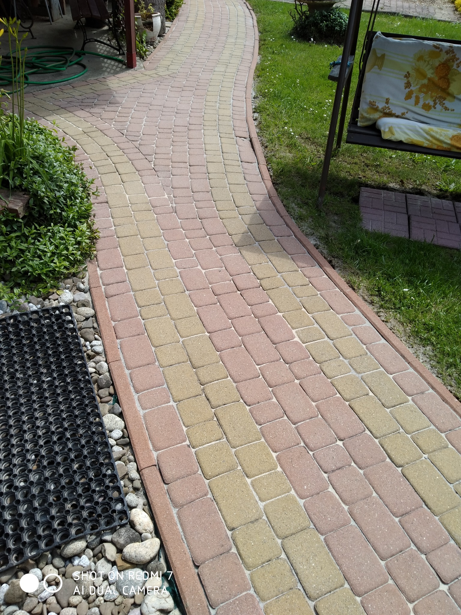Ścieżka z kostki brukowej w odcieniach beżu i różu, wijąca się przez ogród. Widoczny fragment trawnika, rośliny i huśtawka ogrodowa. Kostka czysta, bez zabrudzeń.