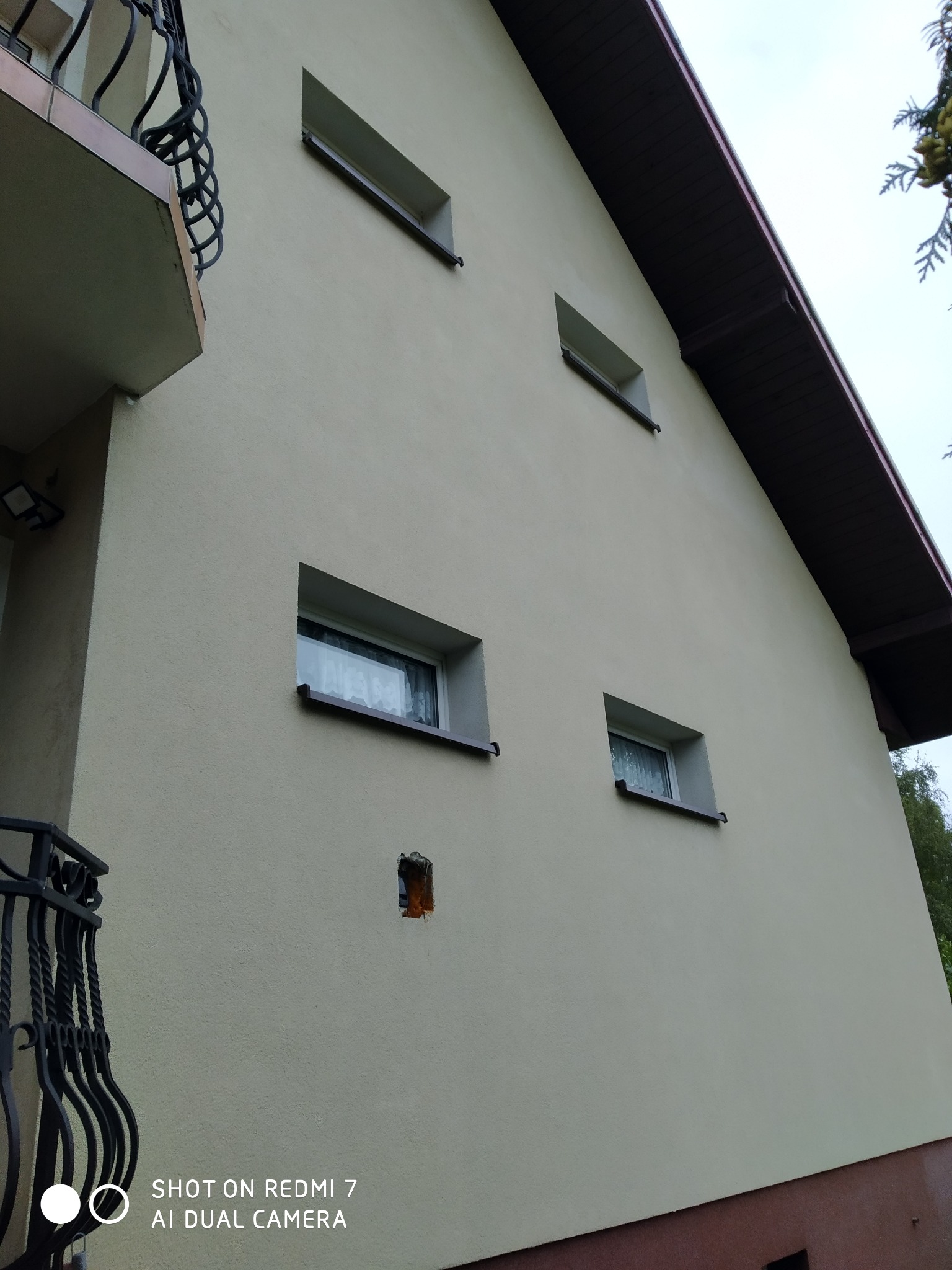 Jasna elewacja domu z widocznymi oknami i fragmentem balkonu z kutą balustradą. Widoczne ślady po usuniętej instalacji na ścianie.
