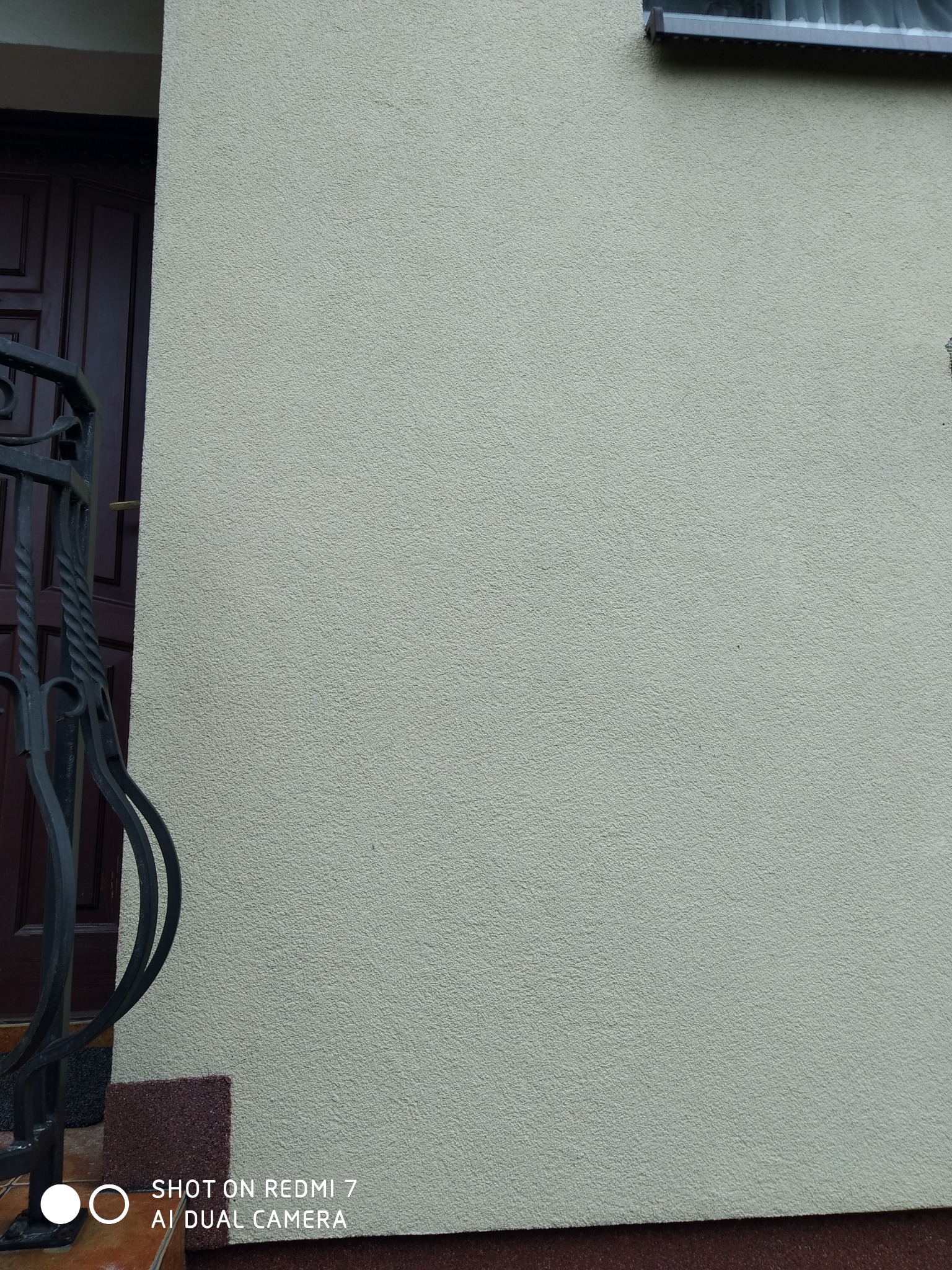 Jasna, ziarnista elewacja budynku z fragmentem okna i ozdobną, ciemną balustradą schodów po lewej stronie.