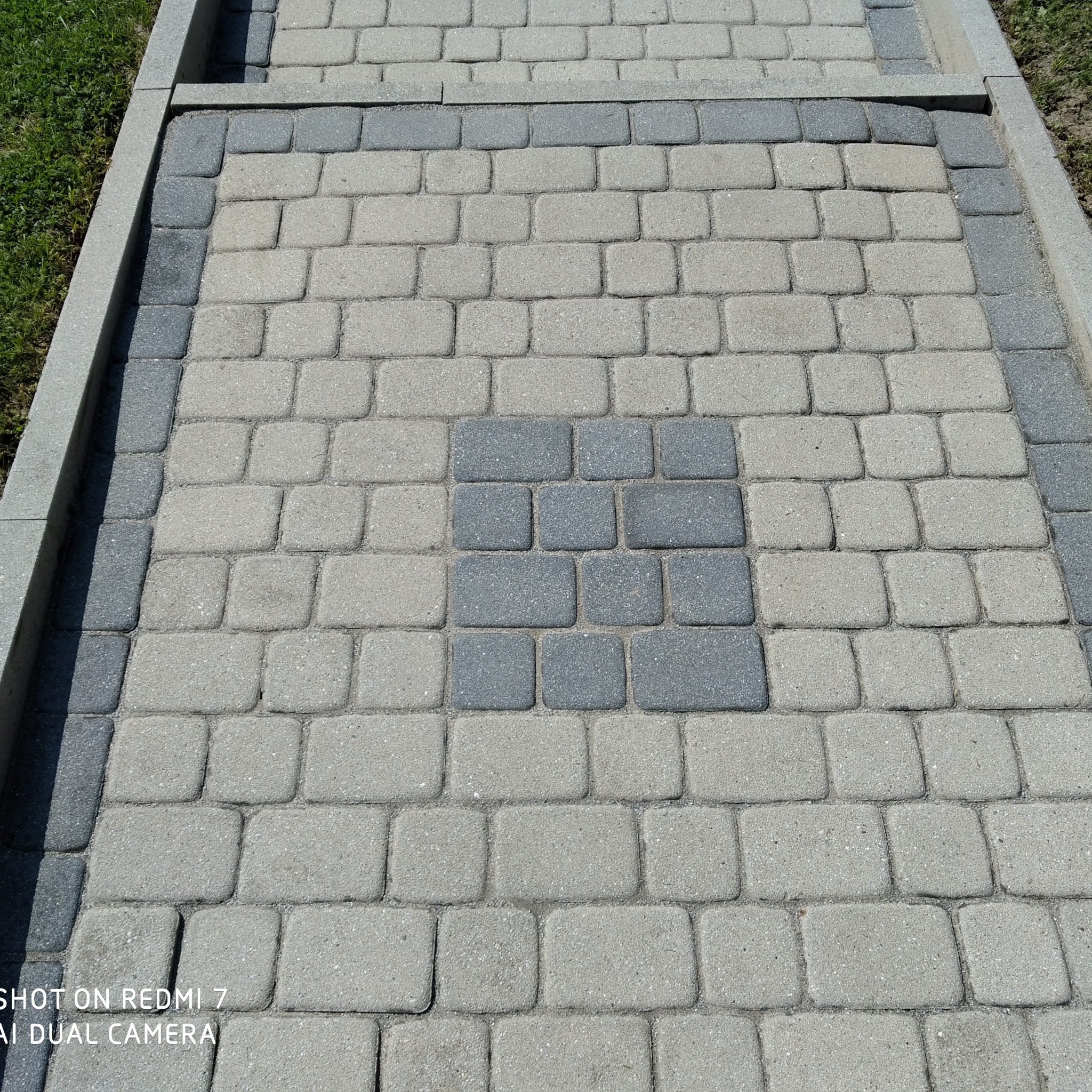 Ułożona kostka brukowa w odcieniach szarości z ciemniejszym kwadratem pośrodku. Widoczne krawężniki i trawa. Zdjęcie z bliska, ukazujące strukturę kostki.