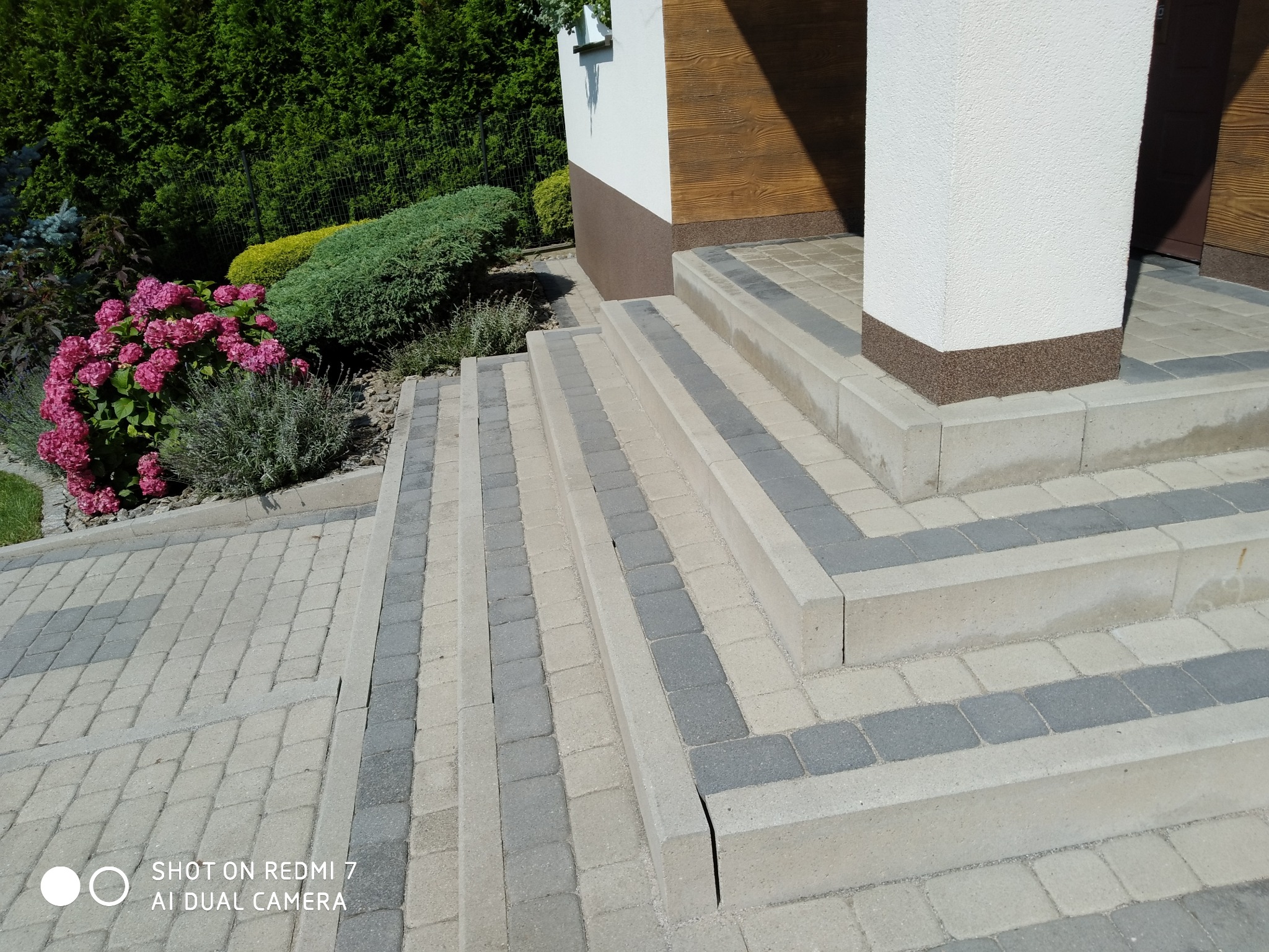 Schody i chodnik z kostki brukowej w odcieniach szarości i beżu, prowadzące do budynku z elementami drewnianymi i kolumną. Obok rosną hortensje.