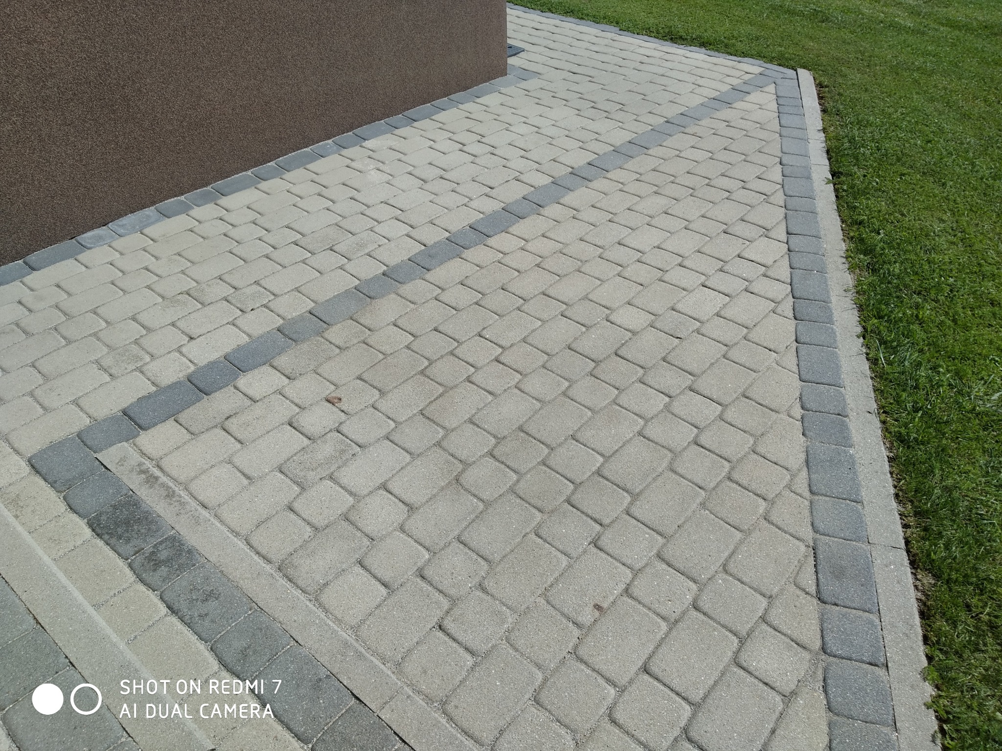 Ułożona kostka brukowa w odcieniach szarości i beżu, z widoczną krawędzią trawnika i fragmentem elewacji budynku. Regularny wzór, świeżo po realizacji.