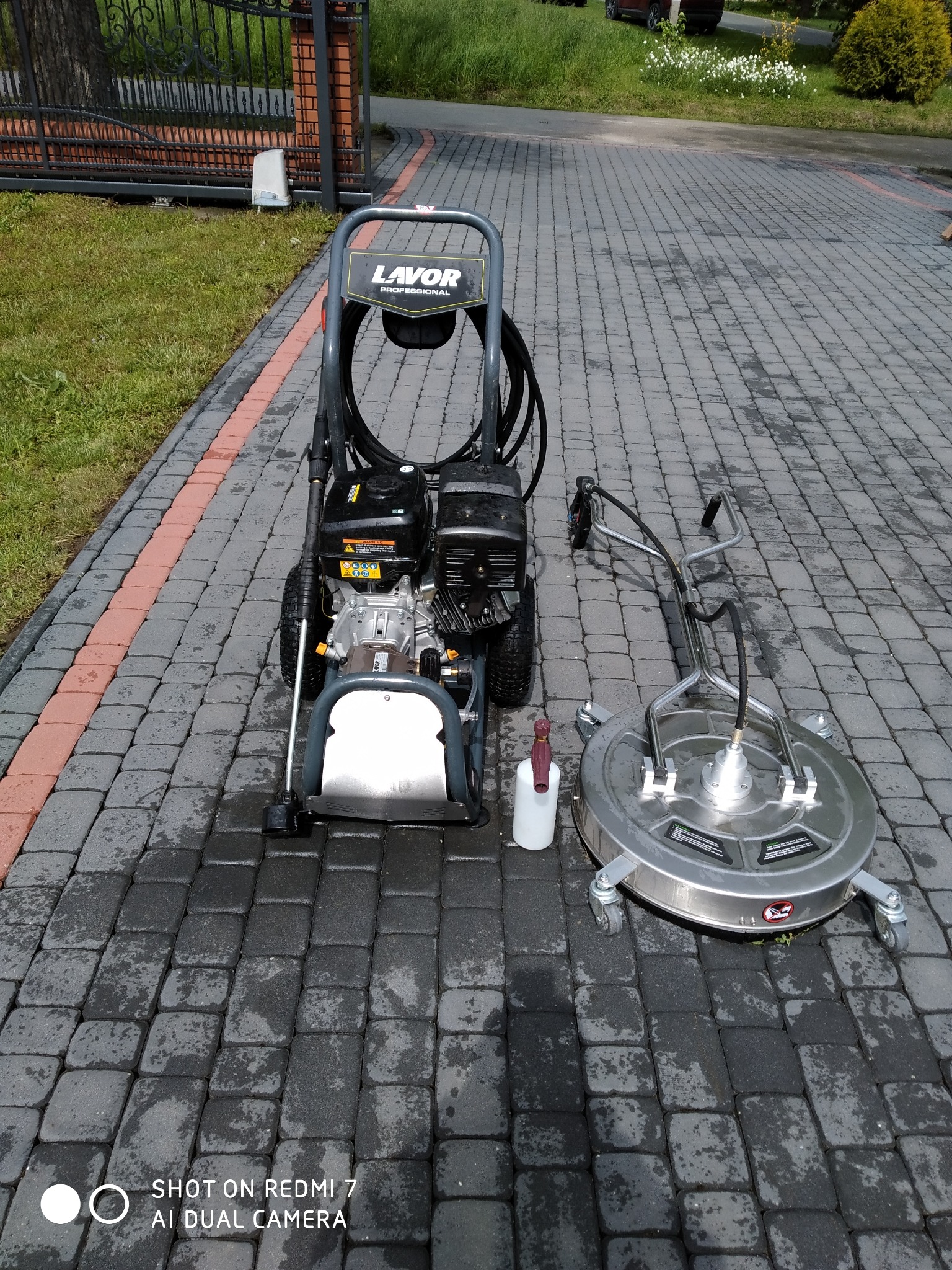 Profesjonalna myjka ciśnieniowa Lavor i okrągła szczotka do kostki brukowej na mokrej, szarej kostce. Widoczne ślady po czyszczeniu i butelka z płynem.