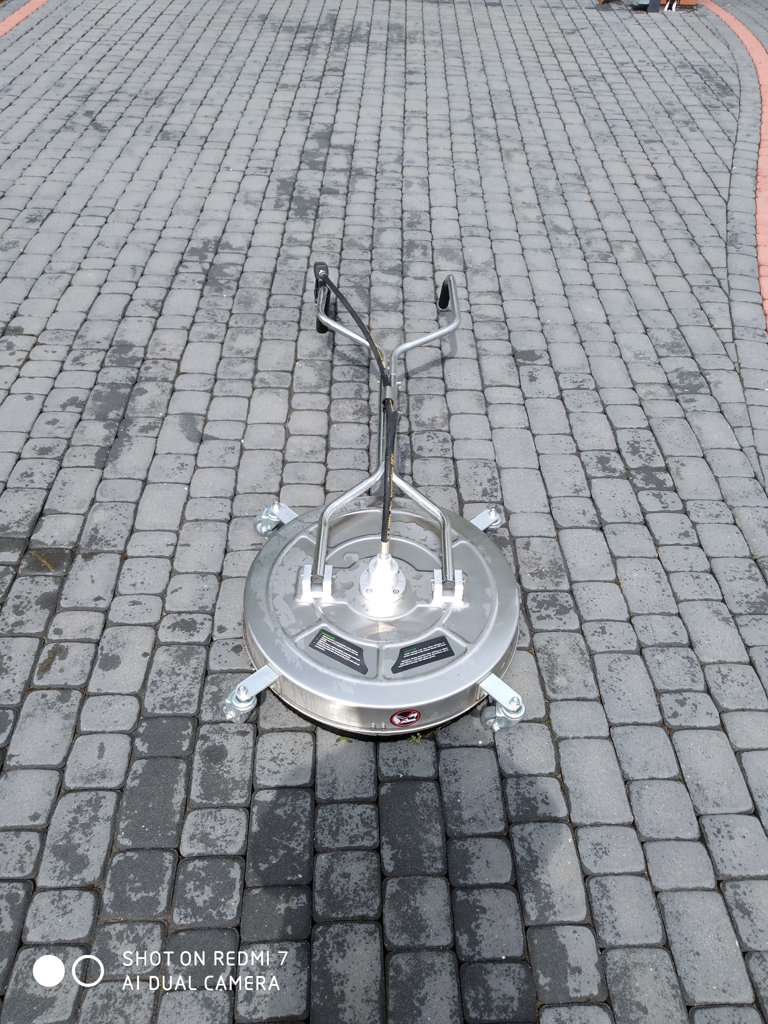 Profesjonalna myjka ciśnieniowa do kostki brukowej, z czterema kółkami, na szarym bruku z delikatnymi zabrudzeniami. Widoczny fragment czerwonego obrzeża.