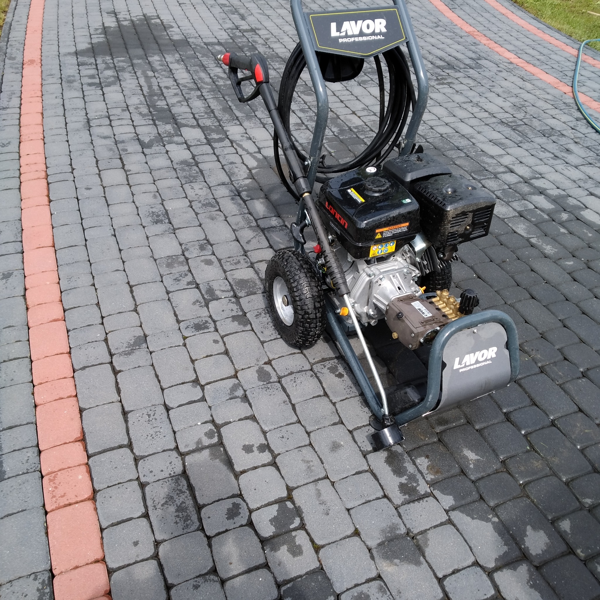 Profesjonalna myjka ciśnieniowa Lavor na kostce brukowej po czyszczeniu, z widocznymi mokrymi plamami. Granitowa kostka z czerwoną obwódką.