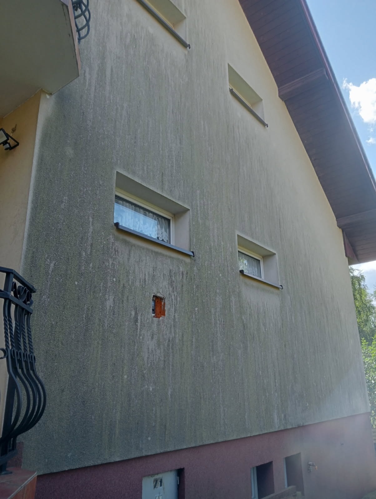 Zabrudzona elewacja dwukondygnacyjnego budynku z widocznymi oknami i fragmentem balkonu. Widoczne ślady zabrudzeń i porostów na ścianie.
