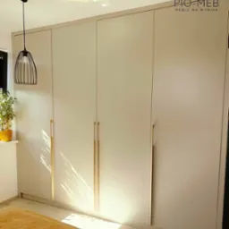 Jasnoszara, czterodrzwiowa szafa z pionowymi, złotymi uchwytami, oświetlona naturalnym światłem, z widocznym fragmentem łóżka w kolorze musztardowym i lampą wiszącą w tle.