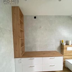 Pio-meb - Białe szafki z drewnianym blatem i regałem w nowoczesnej łazience z szarymi płytkami, oświetleniem punktowym i drewnianą podłogą. Widoczne logo firmy PIO-MEB na górze zdjęcia.