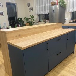 Pio-meb - Nowoczesna kuchnia z drewnianym blatem i szarymi szafkami. Widoczny fragment wyspy kuchennej, okno z firanką i rośliny w tle. Całość w ciepłej tonacji.