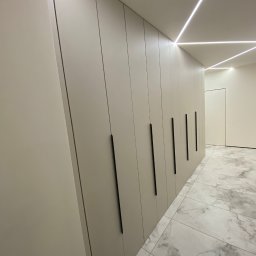 Pio-meb - Długa, zabudowana szafa wnękowa z matowymi, beżowymi frontami i minimalistycznymi, czarnymi uchwytami, oświetlona ledami, w korytarzu z marmurową podłogą.