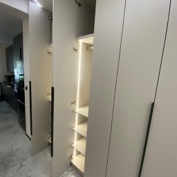 Pio-meb - Nowoczesna szafa wnękowa z oświetleniem LED półek, otwarte drzwi ukazują wnętrze. Minimalistyczny design z długimi, czarnymi uchwytami i jasnymi frontami.