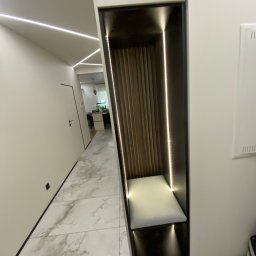 Pio-meb - Nowoczesna zabudowa wnęki z siedziskiem i oświetleniem LED w przedpokoju. Drewniane lamele na ścianie wnęki, marmurowe płytki podłogowe, minimalistyczny design.