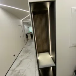 Nowoczesna zabudowa wnęki z siedziskiem i oświetleniem LED w przedpokoju. Drewniane lamele na ścianie wnęki, marmurowe płytki podłogowe, minimalistyczny design.