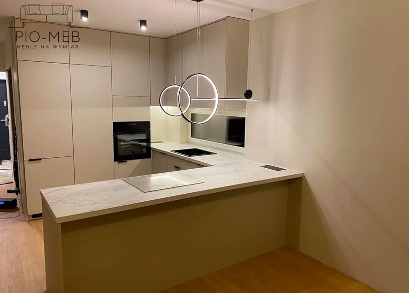 Nowoczesna kuchnia na wymiar z beżowymi frontami, marmurowym blatem i okrągłymi lampami wiszącymi, widoczny piekarnik w zabudowie.