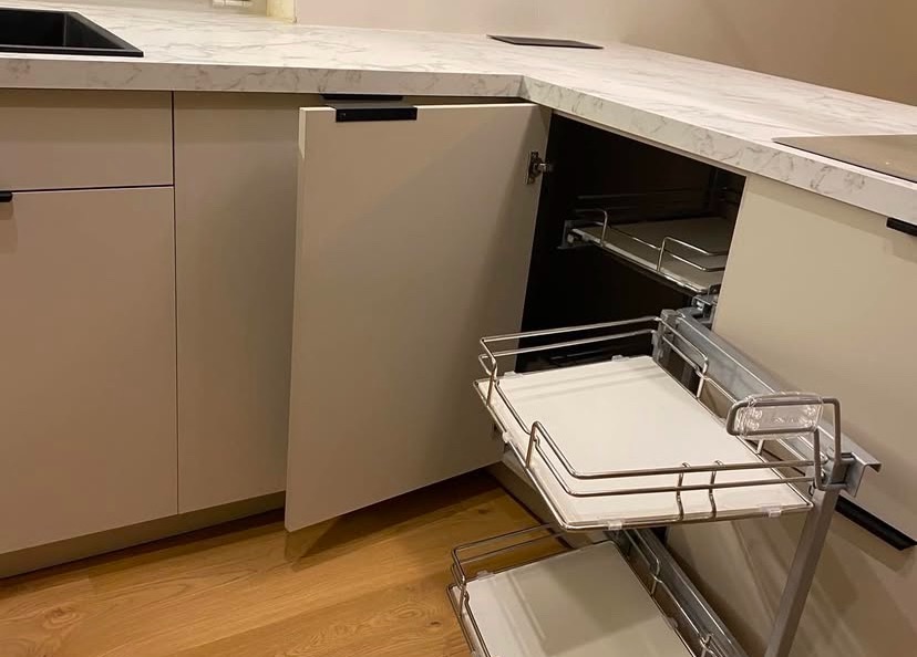 Kremowa szafka narożna z wysuwanymi półkami z metalowym stelażem, widoczna na tle blatu w marmurkowy wzór i drewnianej podłogi.