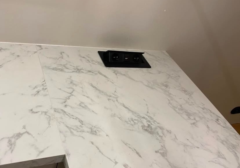 Biały blat z efektem marmuru z wbudowanym, czarnym gniazdkiem elektrycznym.