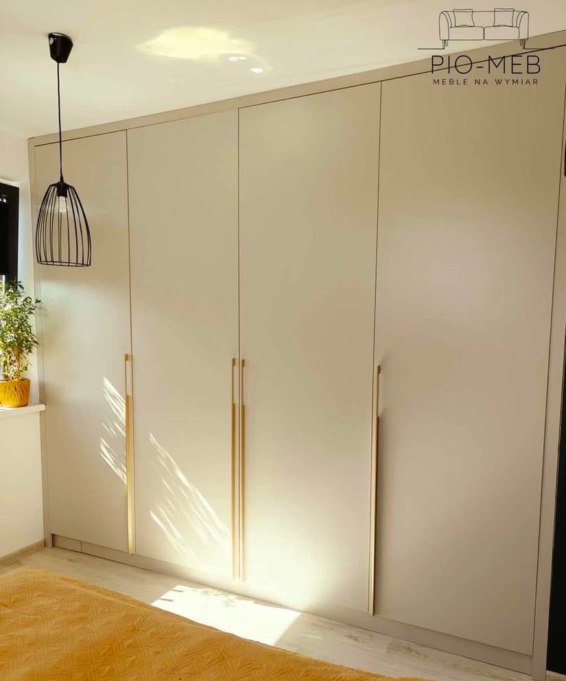 Jasnoszara, czterodrzwiowa szafa z pionowymi, złotymi uchwytami, oświetlona naturalnym światłem, z widocznym fragmentem łóżka w kolorze musztardowym i lampą wiszącą w tle.