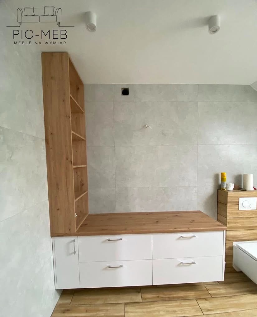 Białe szafki z drewnianym blatem i regałem w nowoczesnej łazience z szarymi płytkami, oświetleniem punktowym i drewnianą podłogą. Widoczne logo firmy PIO-MEB na górze zdjęcia.