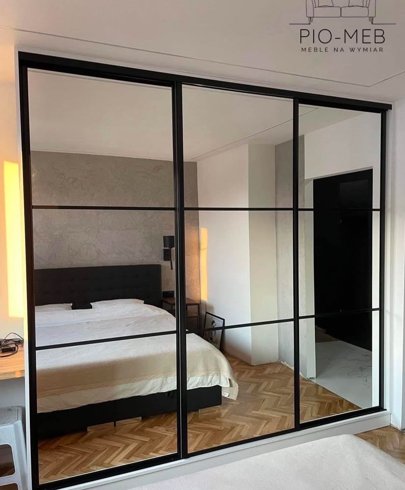 Duża szafa przesuwna z czarnymi ramami i lustrzanymi drzwiami, odbijająca wnętrze sypialni z łóżkiem i parkietem w jodełkę.