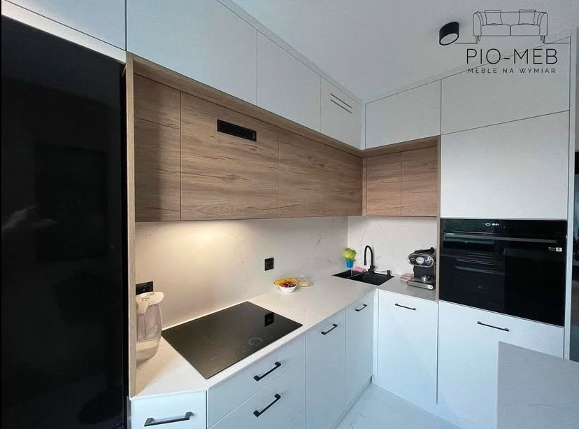 Nowoczesna kuchnia na wymiar z frontami w kolorze drewna i białym blatem, czarne uchwyty, płyta indukcyjna i zabudowany piekarnik, logo firmy Pio-Meb w prawym górnym rogu.