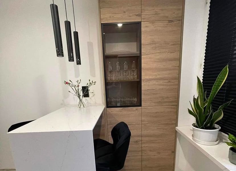 Wysoka zabudowa kuchenna z drewnopodobnymi frontami, przeszklona witryna z kieliszkami, biały blat z szarymi żyłkami, czarne krzesło barowe i lampy tuby