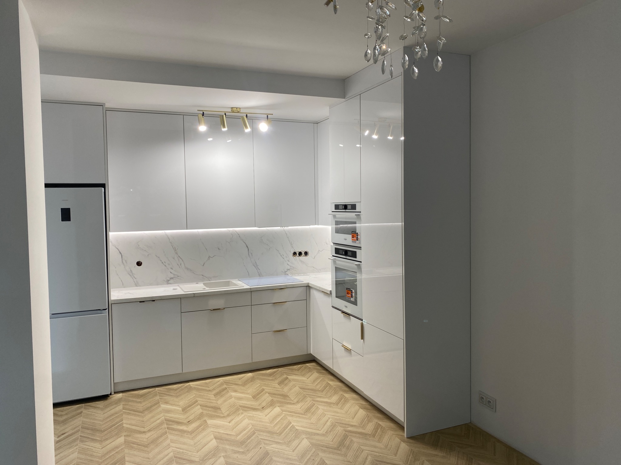 Biała, lśniąca kuchnia z marmurowym blatem i złotymi uchwytami. Nowoczesne oświetlenie sufitowe i parkiet w jodełkę dodają elegancji wnętrzu.