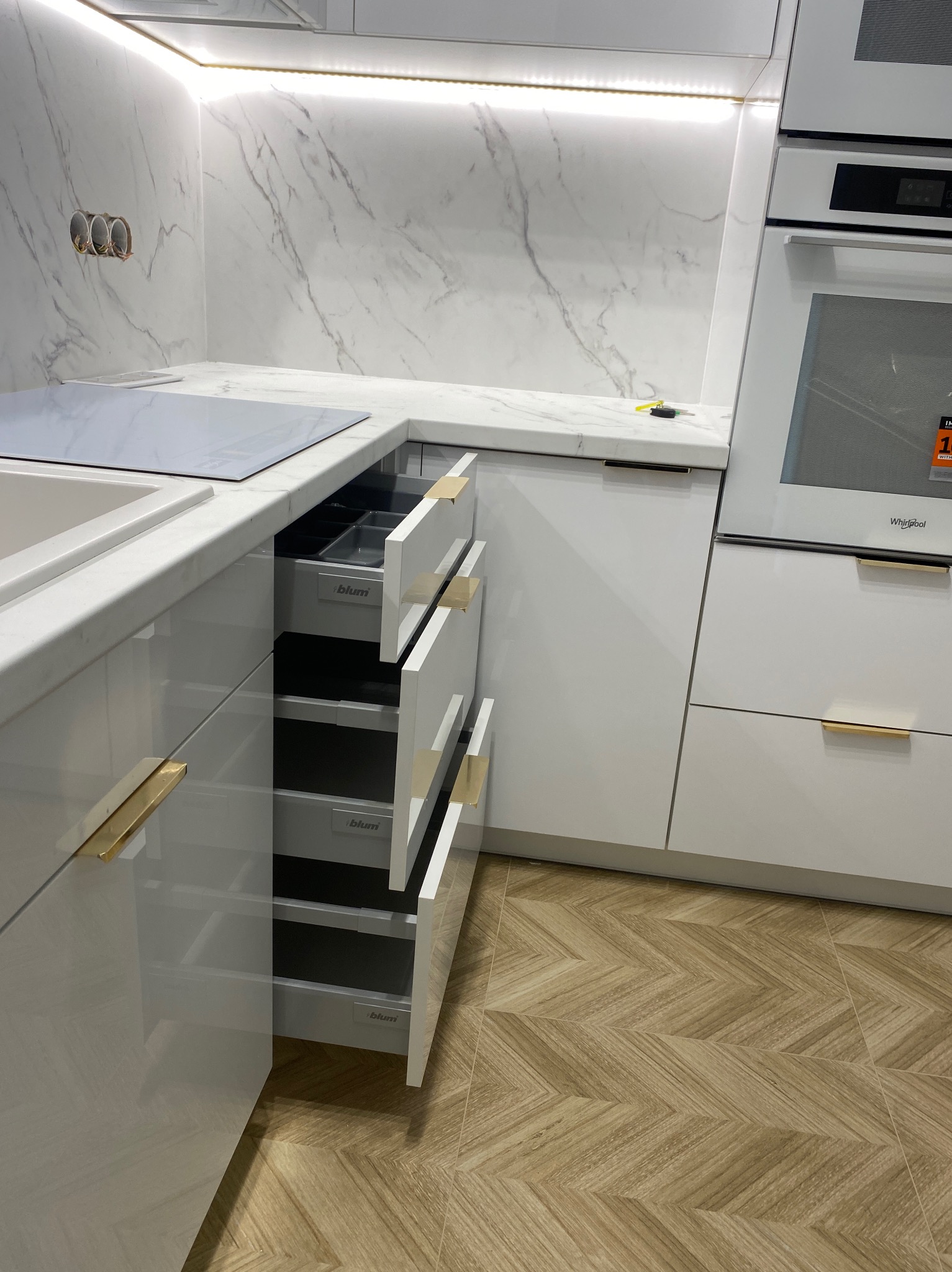 Biała kuchnia z marmurowym blatem, otwartą szufladą Blum i złotymi uchwytami. Widoczne oświetlenie LED podszafkowe i parkiet w jodełkę na podłodze.