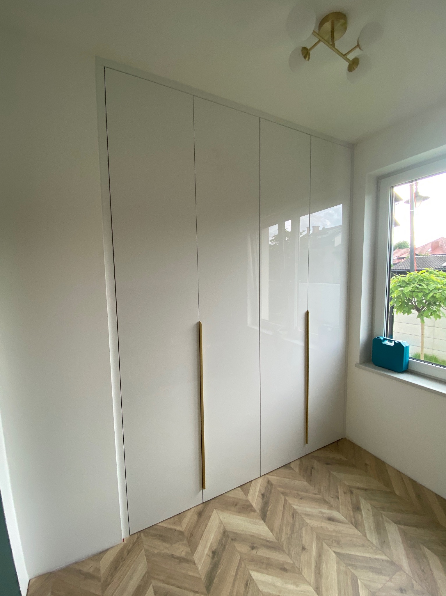 Biała szafa wnękowa z połyskiem, z długimi, złotymi uchwytami. Na parapecie leży turkusowa skrzynka narzędziowa. Parkiet ułożony w jodełkę.