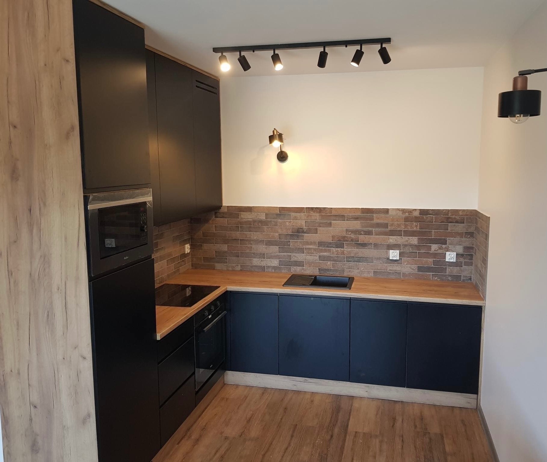 Nowoczesna kuchnia w odcieniach czerni i drewna, z blatem w kolorze dębu i ceglaną ścianą, oświetlona reflektorami sufitowymi i kinkietem.