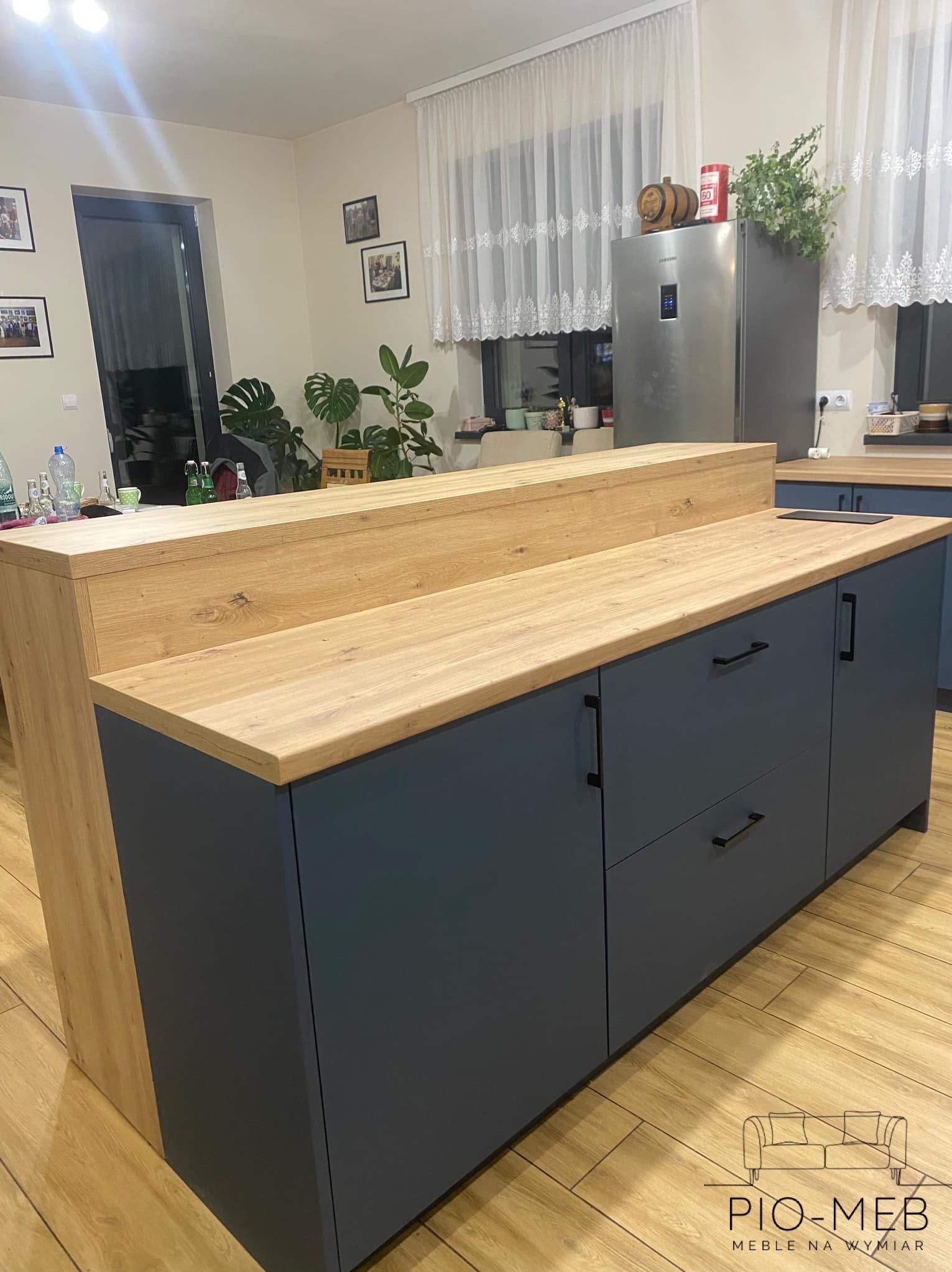 Nowoczesna kuchnia z drewnianym blatem i szarymi szafkami. Widoczny fragment wyspy kuchennej, okno z firanką i rośliny w tle. Całość w ciepłej tonacji.