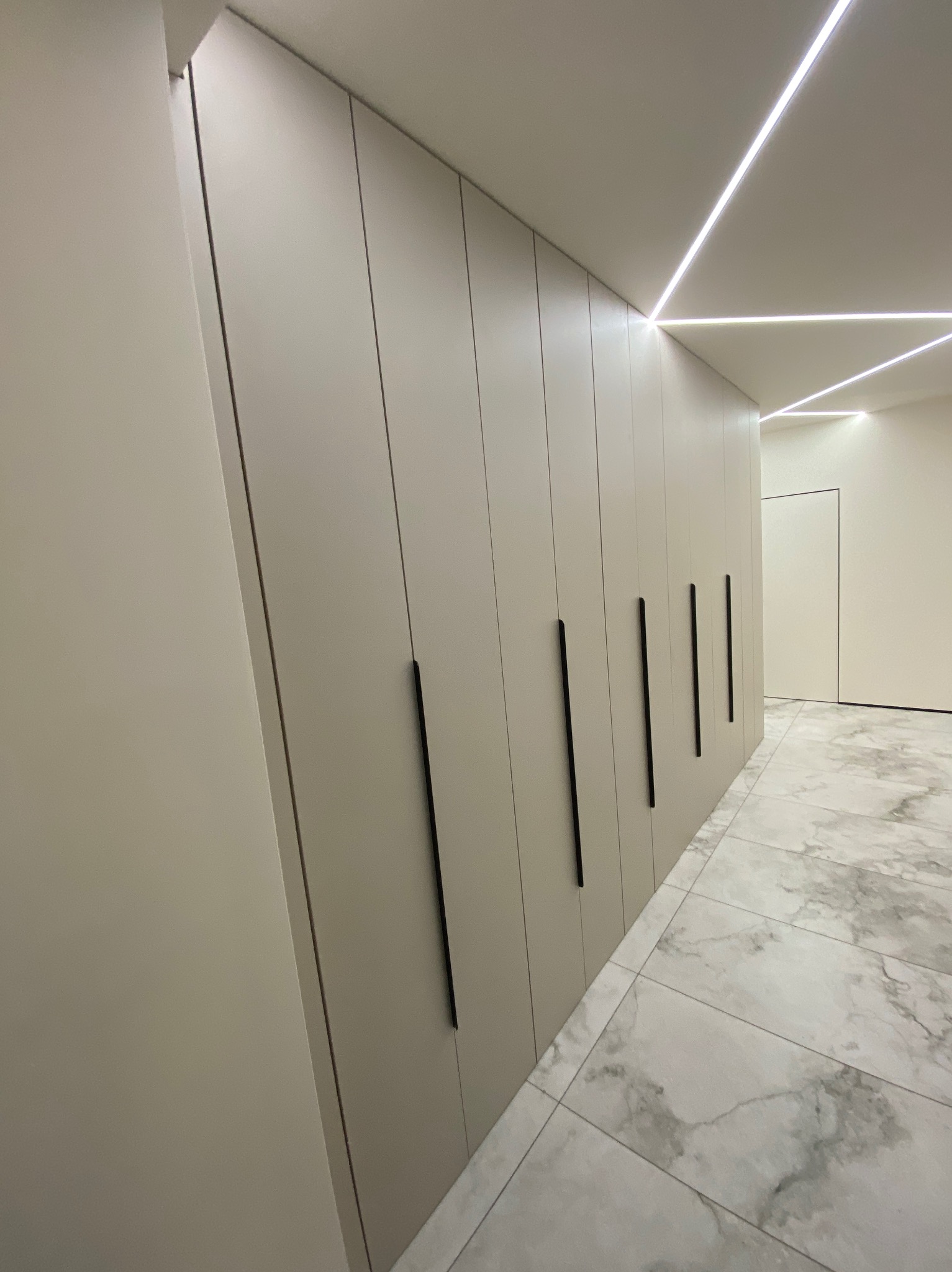 Długa, zabudowana szafa wnękowa z matowymi, beżowymi frontami i minimalistycznymi, czarnymi uchwytami, oświetlona ledami, w korytarzu z marmurową podłogą.